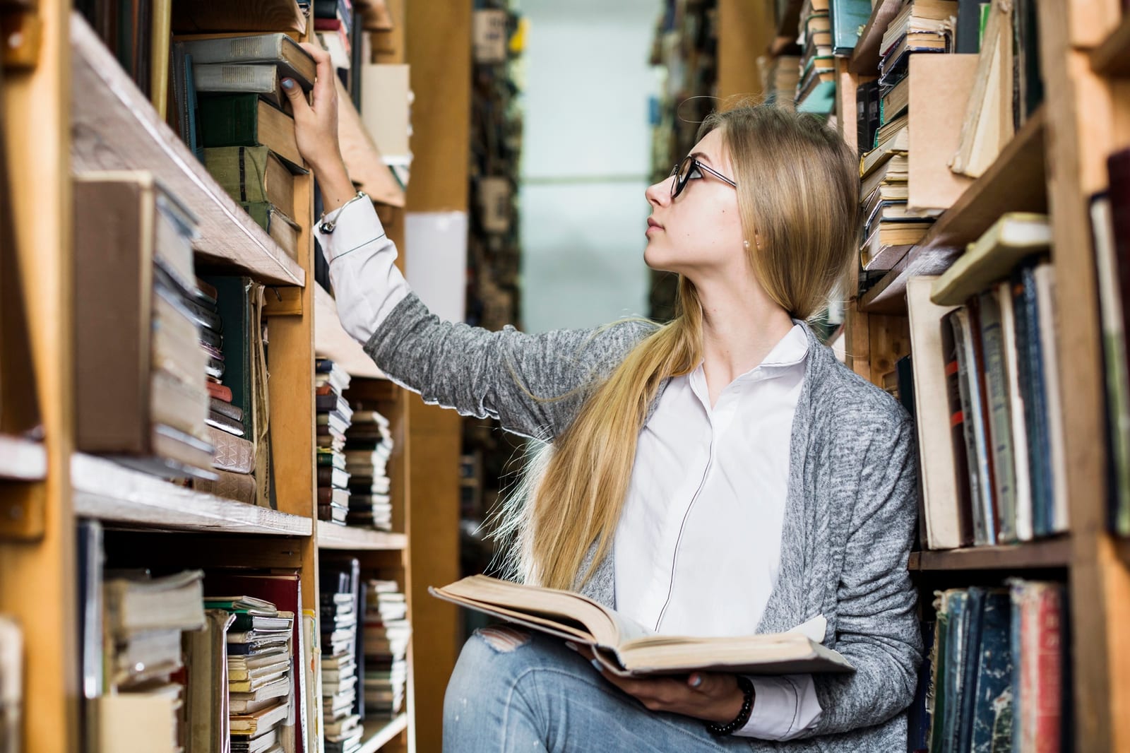 Uma jovem pega um livro em uma estante alta de biblioteca, segurando outro livro aberto no colo.