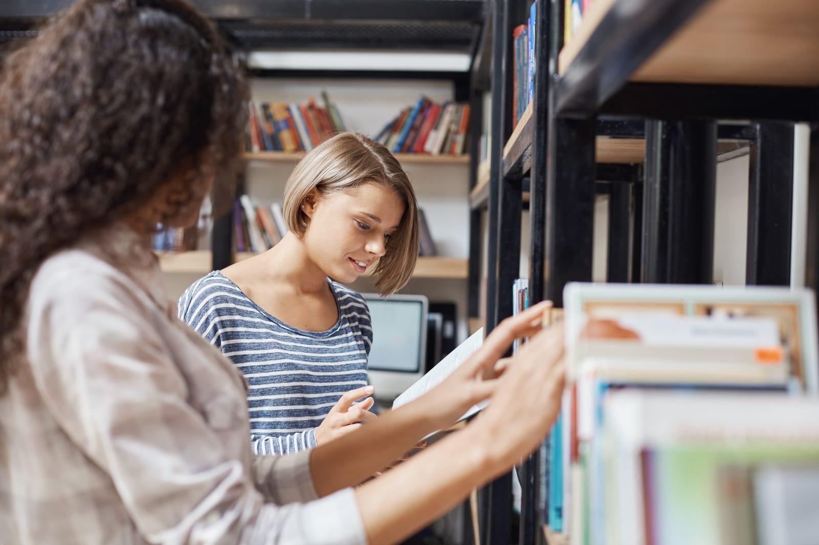 Duas mulheres conversam perto de uma estante de livros, uma delas segurando um livro aberto.