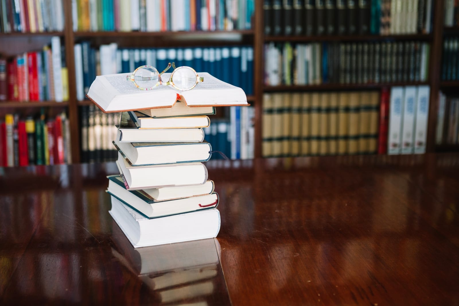 Uma pilha de livros com um óculos de leitura sobre a capa, com estantes de biblioteca desfocadas ao fundo.