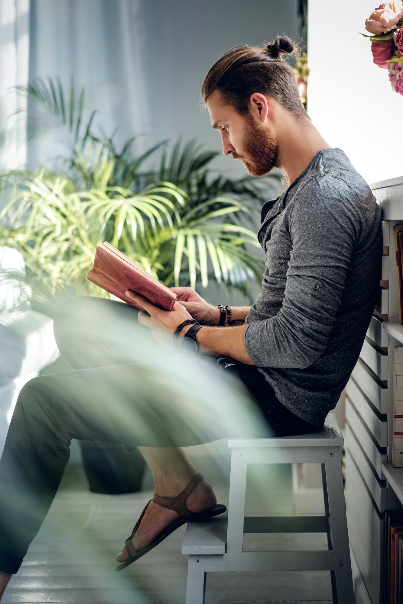 Um homem sentado lê um livro de capa vermelha, com plantas ao fundo.